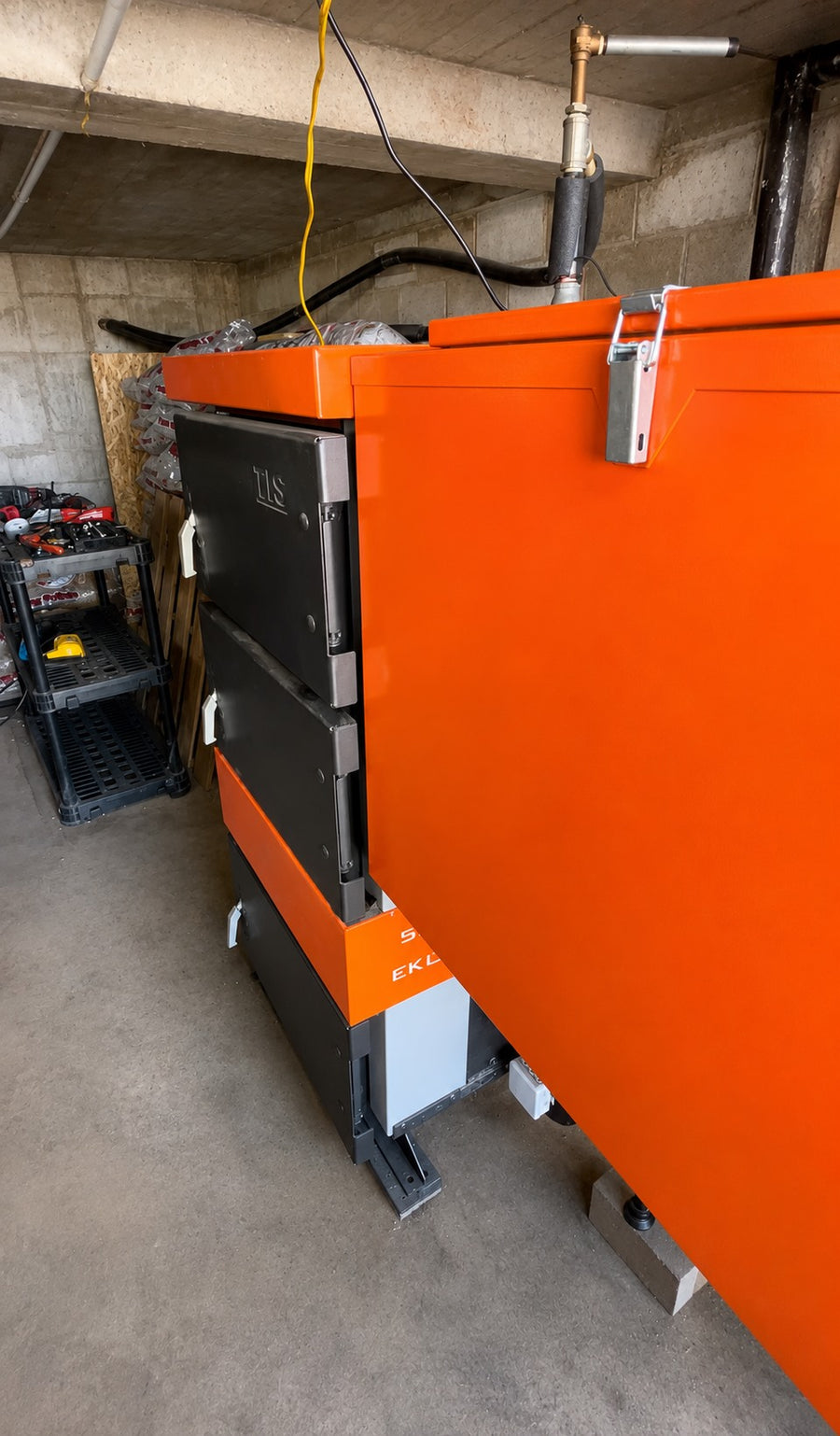 Side view of an orange EKO Duo hybrid coal boiler and integrated fuel hopper in a concrete mechanical room.