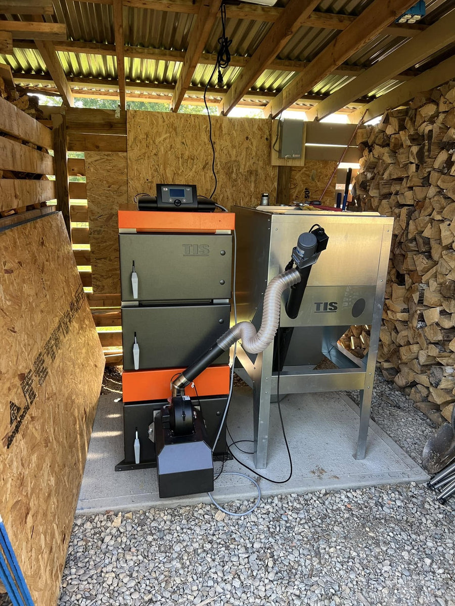 Pellet Duo boiler and metal fuel hopper installed beside stacked firewood in a wood shed.